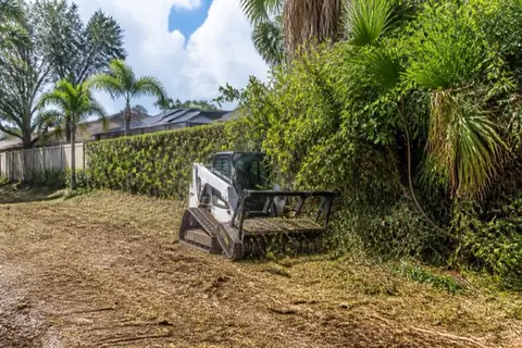 Compact machine clearing shoulder-high brush from the back of a deep Coral Springs lot with a screened pool enclosure and intact ficus hedge beyond the fence