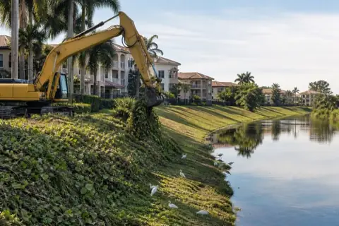 Long-reach excavator removing pepper and vine growth from a residential drainage-canal bank beside a newly mowed clean stretch