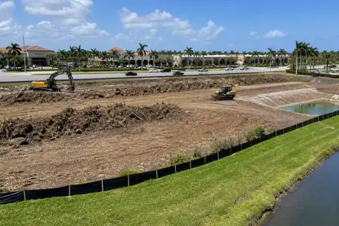 Two unbranded machines clearing mulch windrows inside a silt-fenced commercial outparcel beside a broad Coral Springs arterial under a bright sky