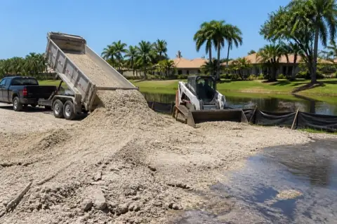 Dump trailer tipping pale sandy fill while a compact skid-steer grades a low lot corner behind silt fence beside a residential canal