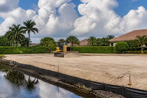 Small dozer spreading pale fill to grade on a Coral Springs homesite with laser level, string lines, preserved trees, and silt fence along the canal-side edge