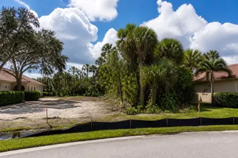 Coral Springs infill lot between occupied stucco homes with one half selectively cleared, survey stakes visible, keeper palms retained, and silt fence protecting the street swale